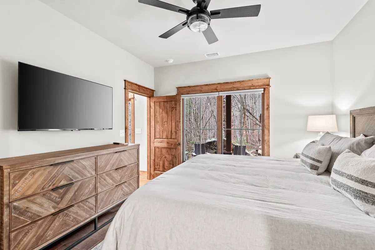 A spacious king suite featuring a large bed with plush bedding, a wooden dresser, a ceiling fan overhead, and a large window showcasing serene nature views. The room is styled in calming, neutral tones to create a relaxing retreat.