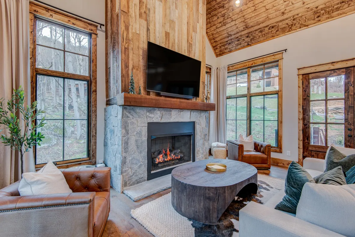 A cozy interior den featuring a gas fireplace as the focal point, surrounded by a large plush sectional and comfortable leather chairs. Large windows let in natural light, and a wooden coffee table sits at the center, inviting gatherings. The space feels spacious yet intimate, perfect for family movie nights or relaxed reading.