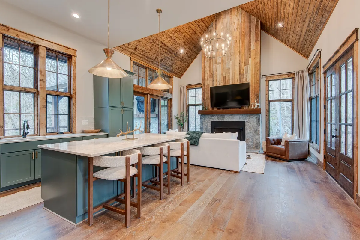 A warm and inviting central living space featuring a kitchen island, stone fireplace, large windows drawing in mountain views, a chandelier above the dining area, and a comfortable sofa. The scene conveys a sense of connection and relaxation, with all major gathering areas visible.