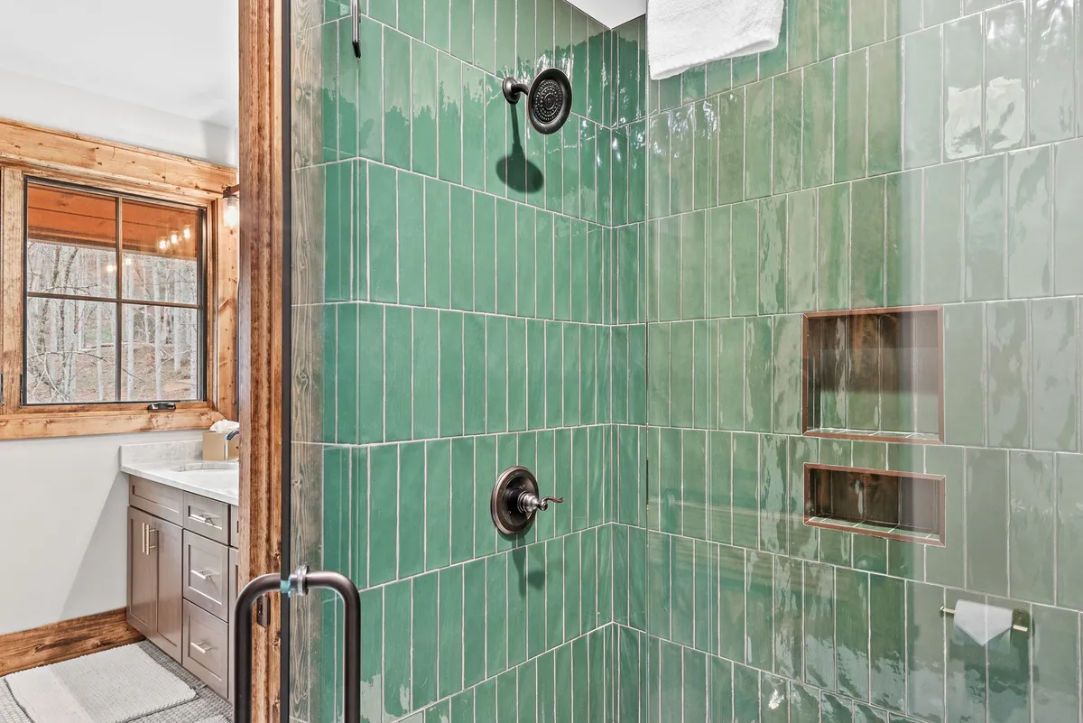 A modern ensuite bathroom with striking green tiled shower walls, black shower fixtures, and a wooden-framed window that lets in natural light. The unique tile choice provides a fresh and stylish look.