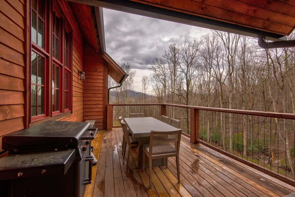 A spacious wooden deck nestled amidst mature trees, featuring a large outdoor dining table set for meals, a modern grill for barbecuing, and panoramic mountain views. The inviting setup offers a seamless blend of comfort and nature, providing ample space for social gatherings or peaceful relaxation.