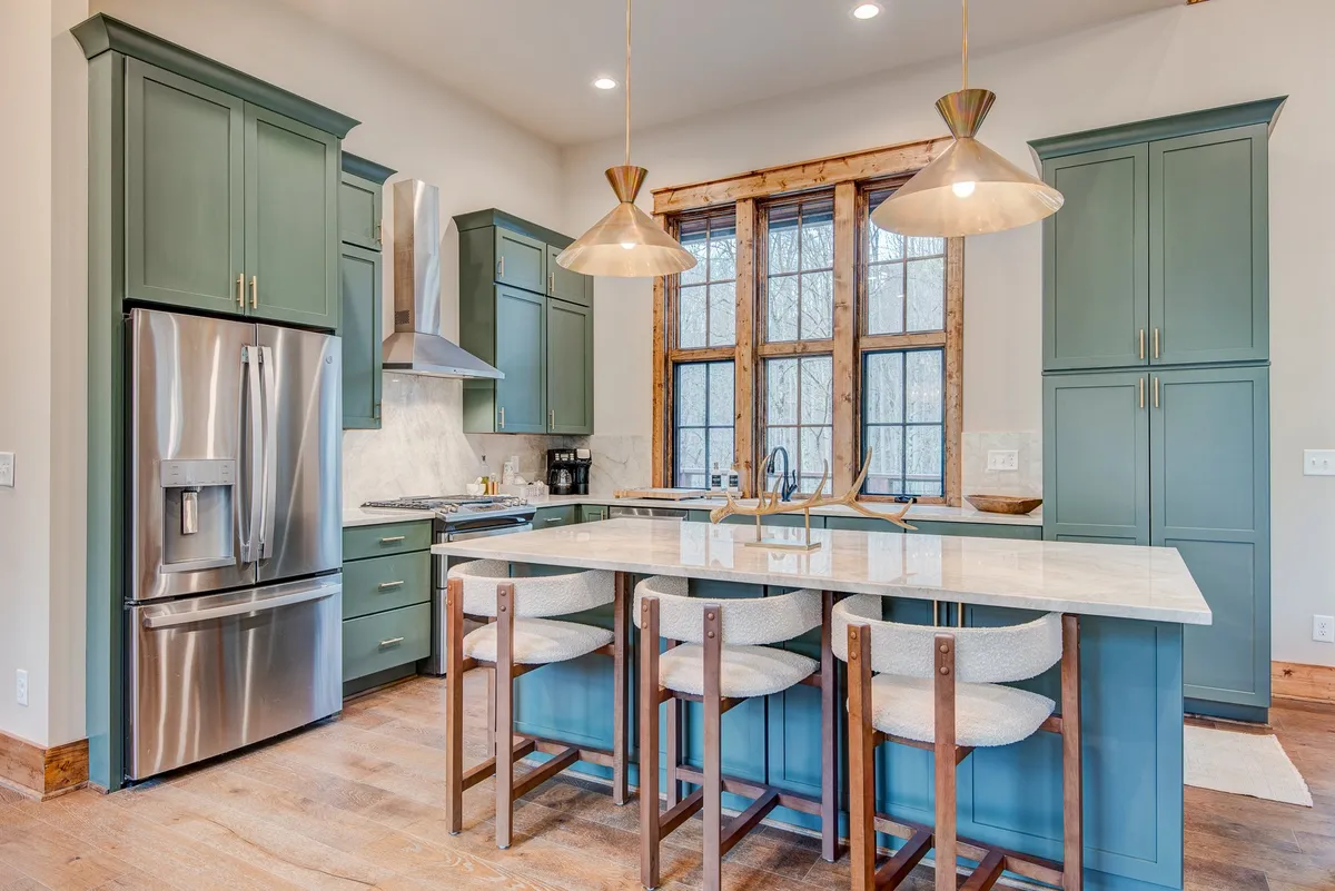 This photo spotlights the kitchen area with an expansive island countertop, large windows framing beautiful views, stylish pendant lights, and cabinetry that adds warmth. The set-up invites both meal preparation and casual conversation, with natural light accentuating the welcoming mood.