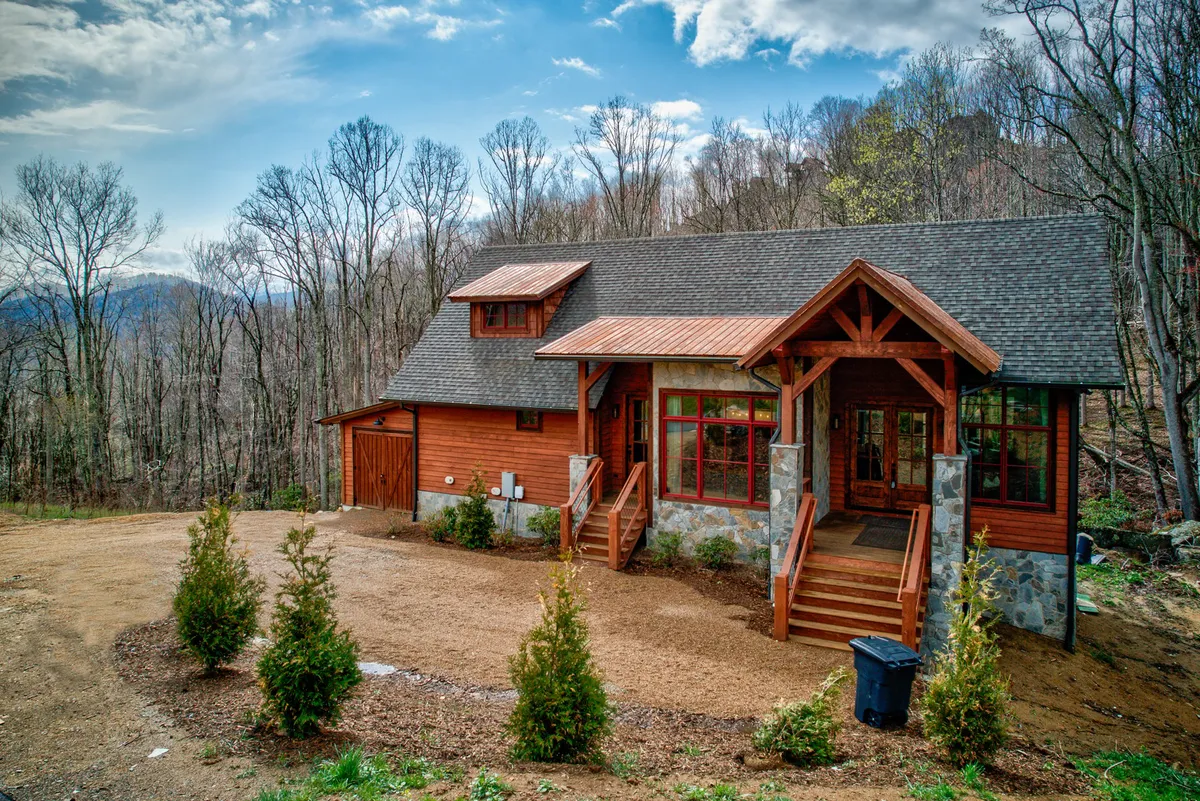 The hero image showcases the inviting exterior of Vesper Lodge, featuring a striking wooden front door framed by elegant stone accents and a classic gabled roof. The cabin is nestled among mature trees, creating a serene, forested backdrop. The spacious porch promises a perfect spot to unwind and take in colorful mountain sunsets.