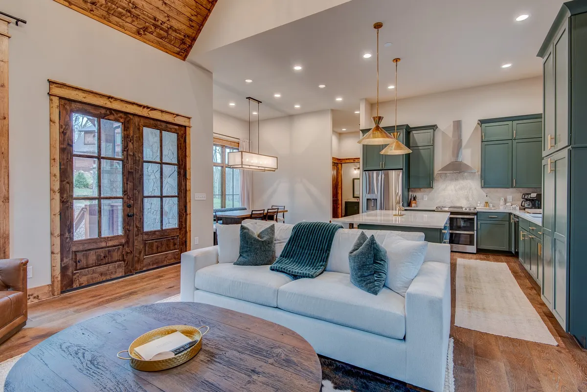 This image showcases an open-concept living and dining space with a cozy sofa adorned with decorative pillows, a prominent wooden dining table, large windows that flood the room with natural light, and a modern kitchen island in the background. The space feels bright, welcoming, and harmoniously connects living, dining, and kitchen areas.