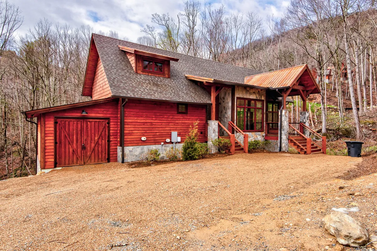 The image features the welcoming front entrance of the lodge, with distinct stone accents, red wooden siding, and a spacious porch with stairs. Surrounded by natural landscaping, the image evokes a sense of privacy and exclusivity in a tranquil mountain setting.