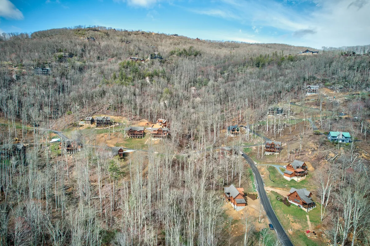 A tranquil mountain community scene showing several wooden cabins nestled along a winding road surrounded by gentle hills and mature trees. The expansive outdoor setting hints at the trails and outdoor activities that begin just steps from the lodge.