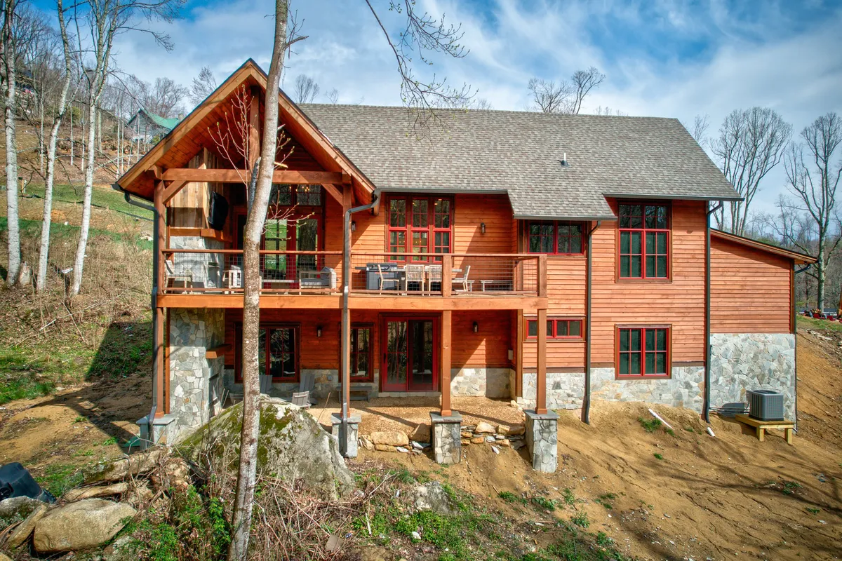 This image shows the exterior of the lodge, featuring a wooden structure with stone foundation, expansive deck area, and large windows that connect indoor and outdoor living spaces. The deck is set up for gatherings, offering views ideal for enjoying sunsets or morning coffee amidst a serene mountain backdrop.